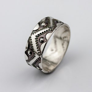 May include: A silver ring with a geometric pattern and small raised dots. The ring is on a white background.