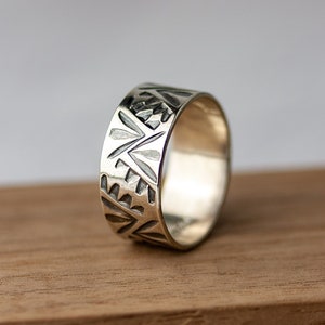 May include: A silver band ring with a geometric pattern. The ring is on a wooden surface.