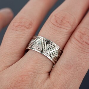 May include: A silver band ring with a geometric design of triangles. The ring is worn on a person's finger.