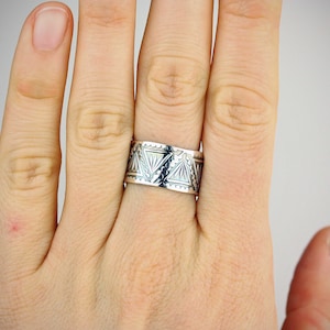 May include: A silver ring with a geometric pattern. The ring is worn on a finger.
