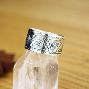 May include: A silver ring with a geometric pattern. The ring is on a clear crystal surface.