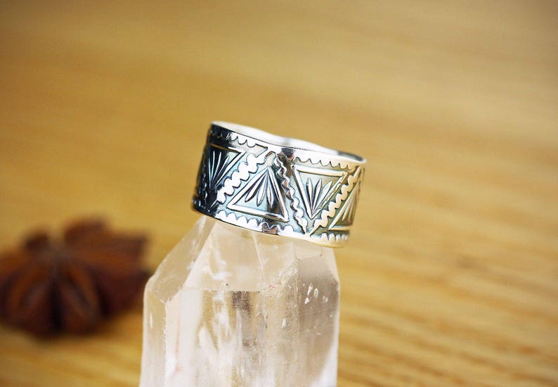 May include: A silver ring with a geometric pattern. The ring is on a clear crystal surface.