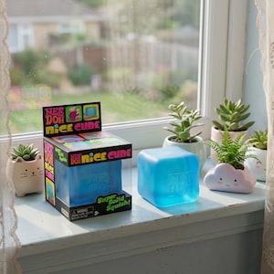 May include: A blue Nee Doh Nice Cube toy and its packaging sit on a windowsill, next to several small potted plants. The cube is a solid, squishy toy. The packaging is black with colorful graphics and the words "Nice Cube".