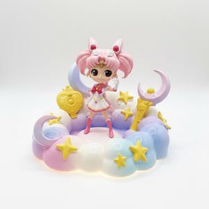 May include: A figurine of a character with pink hair and a white and pink outfit, standing on a pastel-colored cloud base. The base includes yellow stars, a crescent moon, and other decorative elements. The figurine is approximately 5 inches tall.