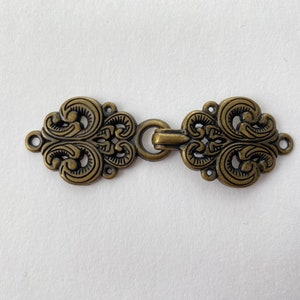 Könnte beinhalten: Ein Paar verzierte, goldfarbene Metallschließen mit komplizierten floralen Designs. Die Schließen sind durch eine einfache Metallscharnier verbunden.