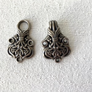 May include: Two ornate silver-toned metal jewelry findings. Each finding has a detailed, symmetrical design with floral and scrollwork elements. One has a loop at the top, and the other has a flat top. These findings are likely for jewelry making.