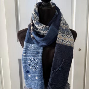May include: A multi-textured scarf featuring various shades of blue knit fabric. One side displays a light blue section with embroidered white floral patterns and small blue buttons, while the other side features a dark blue knit with a pocket detail.
