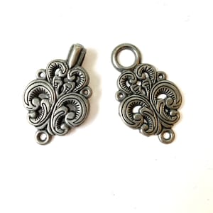 May include: Two ornate, silver-colored clasps with a floral design. One clasp has a loop, and the other has a bar. Each clasp has multiple holes for sewing or attaching to fabric. The clasps are likely for jewelry or garment making.