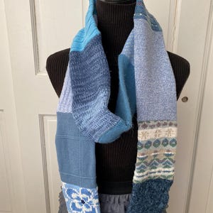 May include: A patchwork scarf in shades of blue, displayed on a black mannequin. The scarf features various knit patterns, including floral and geometric designs. The ends are adorned with decorative buttons, adding a unique touch to this cozy accessory.