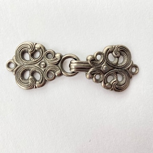 May include: An antique silver-toned clasp with an ornate, symmetrical design. The clasp features a hook and eye closure and is decorated with swirling, scroll-like patterns. The metal hardware is likely used for jewelry or garment closures.