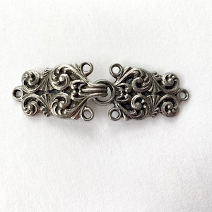 May include: Silver-toned metal clasp with a decorative floral design. The clasp has two loops on each side, allowing for multiple attachment points.