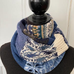 May include: A patchwork infinity scarf in shades of blue, cream, and brown. The scarf features various knit patterns and textures, including a floral cutout design. The scarf is displayed on a black mannequin.