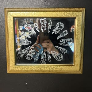 May include: A rectangular mirror framed in textured gold metal, featuring a circular arrangement of white line-art illustrations of various human teeth etched onto the glass surface, creating a repeating pattern around the center of the mirror.