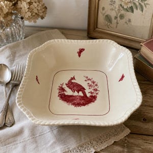 May include: A square cream-colored ceramic bowl featuring a red transferware design of a bird surrounded by floral elements in the center, with small red butterfly motifs on the sides and a textured, scalloped rim detail along the edges.