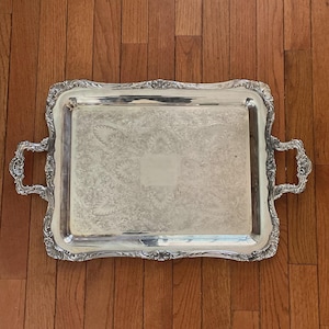 May include: A rectangular silver-toned serving tray featuring ornate, embossed scrollwork along the raised edges and two decorative handles. The central surface displays a detailed, etched floral pattern with a smooth, unadorned rectangular area.