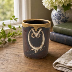 May include: A cylindrical ceramic mug with a dark brown, mottled glaze and a lighter tan rim. The front features a stylized, cream-colored embossed owl design perched on a branch, with circular eyes and a pointed beak, creating a textured relief.