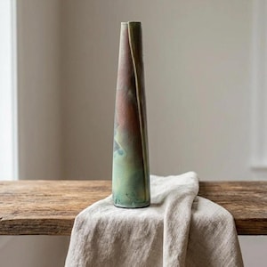May include: A tall, slender, conical ceramic vase featuring a textured surface with a gradient of muted green, brown, and charcoal tones. The vase has a narrow opening at the top and a vertical seam running down its side, showcasing a matte finish.