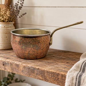 May include: A round copper saucepan with a weathered, textured exterior and a metallic patina. The pot features a long, slender brass handle attached to the side with a single rivet and a smooth, light-colored interior rim.