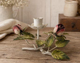 May include: A metal candle holder featuring a central white cup-shaped candle seat surrounded by two sculpted rose buds with red and white petals, accented by green leaves and white-painted branches, designed to hold a single taper candle.