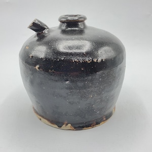 May include: A dark brown, glazed ceramic jug with a rounded body, a central top opening, and a small side spout. The surface features a textured, glossy finish with visible imperfections and a lighter, unglazed base rim showing the clay body.