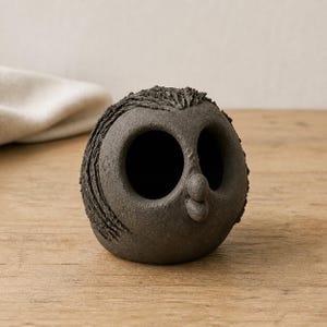 May include: A dark charcoal gray ceramic sculpture shaped like a round owl with a textured, rough surface. The figure features two large, deep, hollow circular eye sockets and a small, protruding, rounded beak centered between the eyes.
