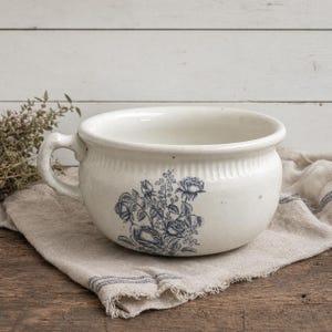 May include: A cream-colored ceramic chamber pot featuring a single side handle and a blue floral transferware pattern on the front. The upper rim of the vessel is decorated with a subtle vertical ribbed texture, and the interior is smooth.