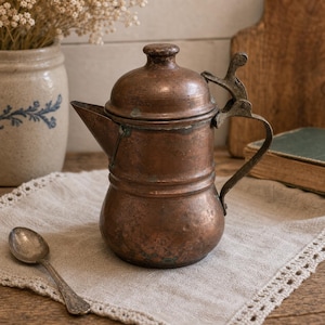 May include: A weathered copper coffee pot with a rounded body, a short spout, and a hinged lid featuring a metal thumb lever. The surface shows a dark, aged patina with visible oxidation spots. A small, tarnished silver-toned metal spoon lies nearby.