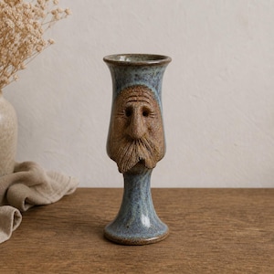 May include: A tall ceramic goblet with a blue and grey speckled glaze. The front of the vessel features a sculpted, textured brown face with a prominent nose, deep-set eyes, and a detailed mustache, all integrated into the stem and bowl design.
