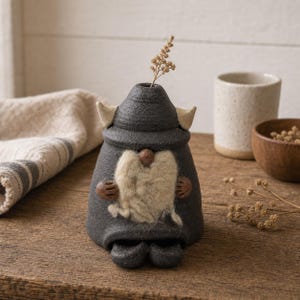 May include: A dark gray ceramic gnome figurine with a textured finish, featuring a cream-colored felted beard, two small horns on the sides of its hat, and a small brown nose. A single dried plant stem is inserted into the top of the gnome's hat.