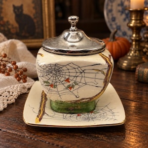 May include: A cream-colored ceramic sugar bowl with a matching square saucer and a silver-toned metal lid. The bowl and saucer feature a hand-painted design of black spiderwebs, green grass, and brown tree branches with small orange accents.