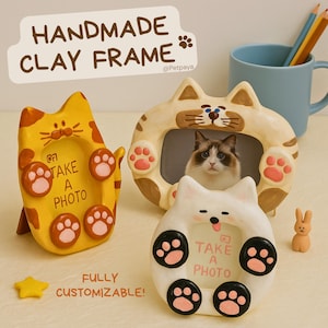 May include: Handmade clay cat-shaped photo frames in yellow, cream, and white. Each frame has paw prints and the text "TAKE A PHOTO." The frames are customizable and have a cartoonish design. A blue mug and pencils are in the background.