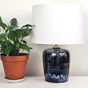 May include: A navy blue ceramic table lamp with a white lampshade. The lamp has a glossy finish with a white and blue pattern at the base. A potted plant with green leaves sits to the left.