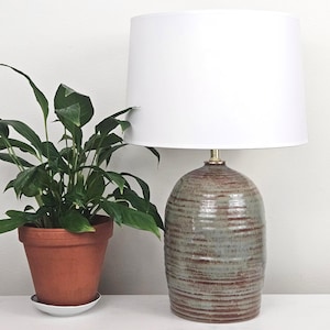 May include: A ceramic table lamp with a white lampshade. The lamp base is a textured, earthy green and brown. A potted plant with green leaves sits to the left of the lamp. A framed print hangs above the lamp.