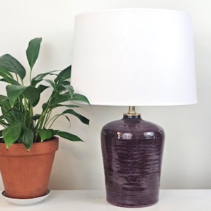 May include: A decorative table lamp with a white lampshade and a dark purple, textured ceramic base. The lamp sits on a white surface next to a potted green plant. The lamp has a brass-colored neck.