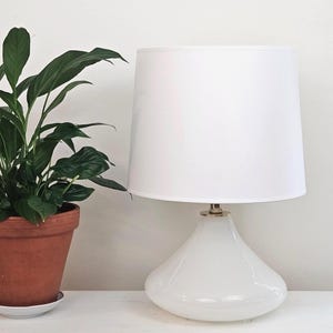May include: A white table lamp with a rounded base and a white lampshade. The lamp is next to a potted plant with green leaves in a terracotta pot. The plant sits on a white surface against a white wall.