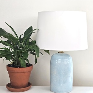 May include: A table lamp with a cylindrical, light blue ceramic base featuring a subtle crackle glaze finish and a brass-toned neck, topped with a large, plain white drum lampshade. The lamp stands next to a green plant in a terracotta pot.