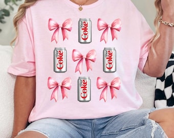 Op de afbeelding: Een lichtroze t-shirt met een patroon van roze strikken en Diet Coke-blikjes. De Diet Coke-blikjes zijn zilverkleurig met rode en witte tekst. De strikken zijn zachtroze.
