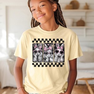 May include: A pale yellow t-shirt with a graphic of three bunnies wearing heart-shaped sunglasses and pink bows. The graphic is framed by a black and white checkered border. The shirt is being worn by a person.