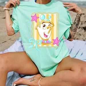 May include: A pale green t-shirt with a cartoon teacup character design. The design includes a yellow and white striped background, pink starfish, and blue bubbles. The teacup has a smiling face and a blue and pink collar. The shirt is worn on a beach.