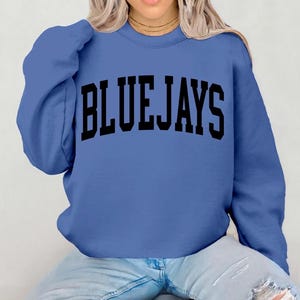 Peut inclure: Un sweat-shirt bleu pervenche avec l'inscription "BLUE JAYS" en grosses lettres noires. Le sweat-shirt est de style col rond et semble être fait d'une matière douce.