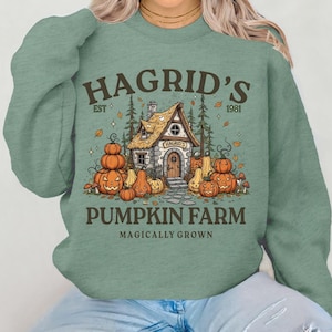 Peut inclure: Sweat-shirt vert sauge avec un motif de maison en pierre entourée de citrouilles et d'arbres. Le texte indique "Hagrid's Pumpkin Farm" et "Magically Grown". Le design comprend le texte "Hagrid's" et "Est 1981".