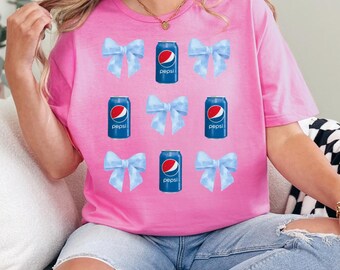 Op de afbeelding: Een roze t-shirt met een patroon van blauwe strikken en Pepsi-blikjes. De Pepsi-blikjes zijn blauw met het Pepsi-logo. De strikken zijn lichtblauw. Het shirt is een crewneck met korte mouwen.