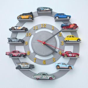 May include: A gray wall clock with a circular design, featuring miniature toy cars arranged around the clock face. The clock hands are black with a red second hand, and the numbers are gold. The cars are various colors.