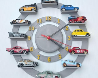 May include: A gray wall clock with a circular design, featuring miniature toy cars arranged around the clock face. The clock hands are black with a red second hand, and the numbers are gold. The cars are various colors.