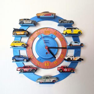 May include: A circular blue wall clock with a speedometer-inspired orange face featuring numbers from 10 to 120. The clock frame has ten small shelves, each holding a miniature toy car. The centre includes black clock hands and the text KM and FUEL.
