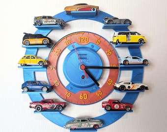 May include: A circular blue wall clock with a speedometer-inspired orange face featuring numbers from 10 to 120. The clock frame has ten small shelves, each holding a miniature toy car. The center includes black clock hands and the text KM and FUEL.