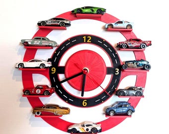 May include: A circular wall clock featuring a red frame with twelve small shelves, each holding a miniature toy car. The clock face has a black track design with yellow numbers 3, 6, 9, and 12, and black hands. The cars vary in color and style.