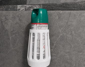 May include: A white plastic wall-mounted holder containing an aerosol spray can with a green cap. The holder features a series of vertical slats that reveal the label of the spray can, which includes printed text and a red hazard warning symbol.