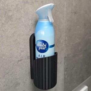 以下が含まれることがあります： 垂直にリブが入った黒い壁掛けホルダーに、水色のAmbi Pur Airスプレーボトルが収められています。ホルダーは湾曲したデザインでエアゾール缶を固定しており、缶には白いトリガーノズルとブランドロゴが印刷されています。