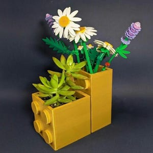 以下が含まれることがあります： 2段になった黄色のプラスチック製ブロックのプランター。緑色の多肉植物と、黄色い中心を持つ白いデイジーやブロックで作られた紫色のラベンダーのような茎を含む造花のブーケが飾られています。